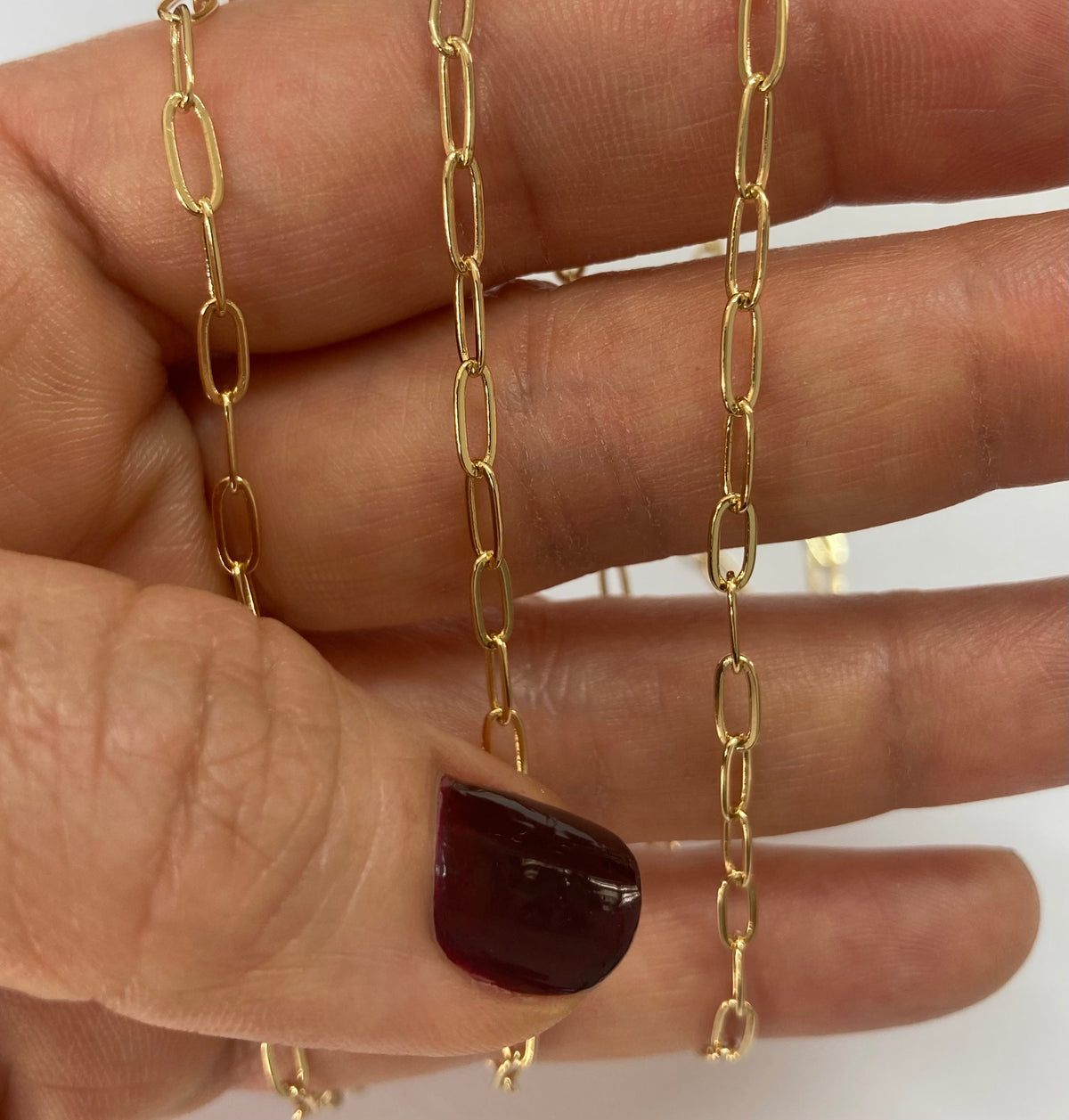 A close-up of a hand holding two gold paperclip chain necklaces against a plain white background.