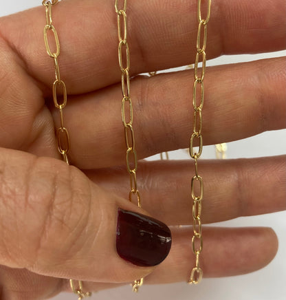 A close-up of a hand holding two gold paperclip chain necklaces against a plain white background.