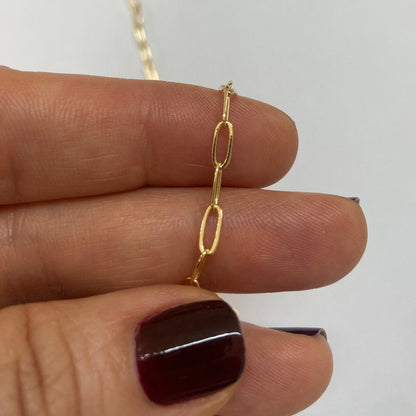A close-up of a delicate gold paperclip chain necklace held between fingers against a plain light background.