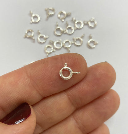 A close-up of a person's hand holding a small sterling silver ring clasp, with several matching silver ring clasp pieces scattered in the background on a white surface.