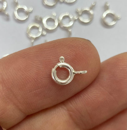 A close-up of a small silver spring ring clasp held between fingers, with several similar clasp pieces scattered in the background on a white surface.